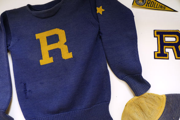 A collection of Rollins memorabilia, including a tattered blue knit sweater with an embroidered gold "R", a small gold pennant with the text "Rollins", a blue "R" patch with a gold edge, and a used beige fishing hat with a blue brim