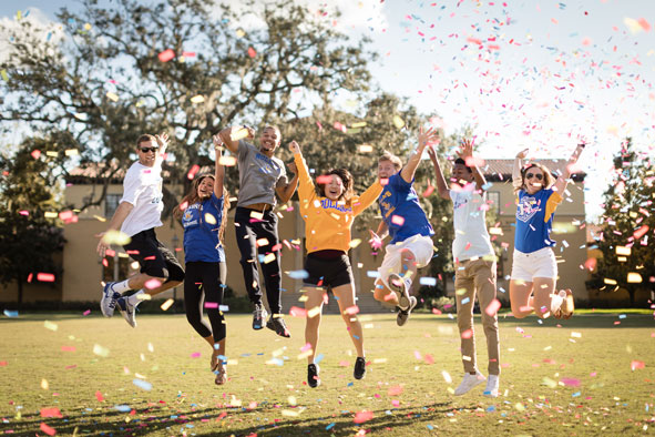 Seven young students jumping high in the air with arms outstretched with confetti thrown in the air on an open lawn